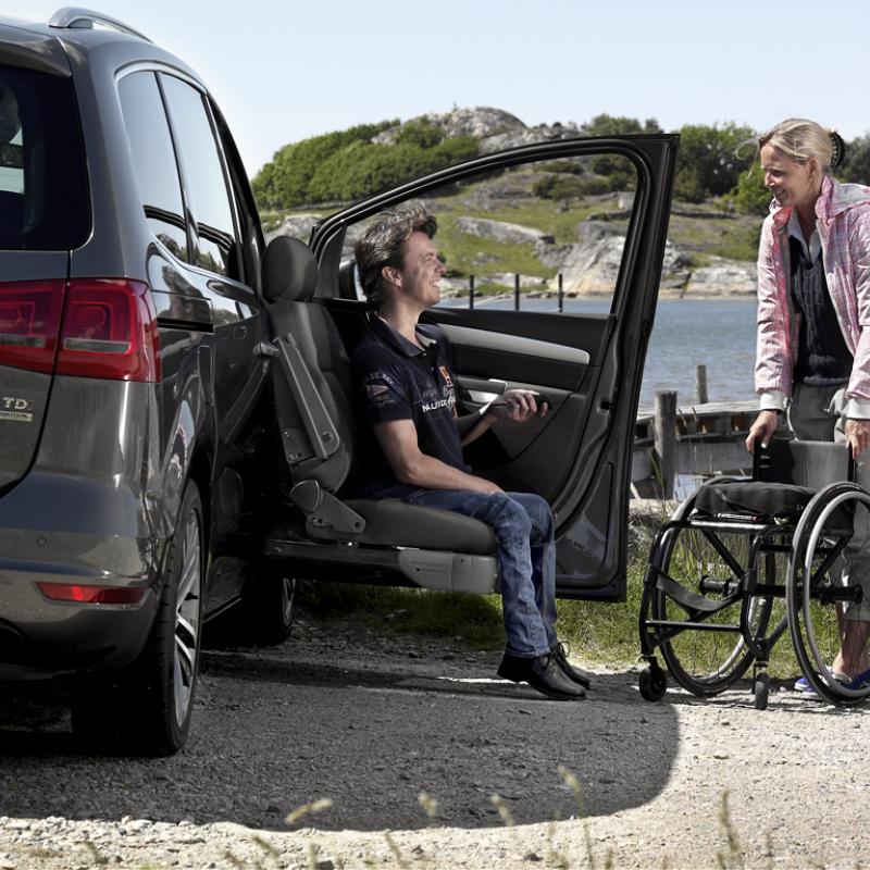 Carer brings a wheelchair to a user sitting in a seat lift of a car. 