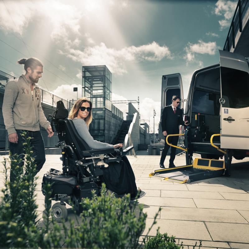User in powerchair with her carer moving towards a minibus with a deployed wheelchair lift. 