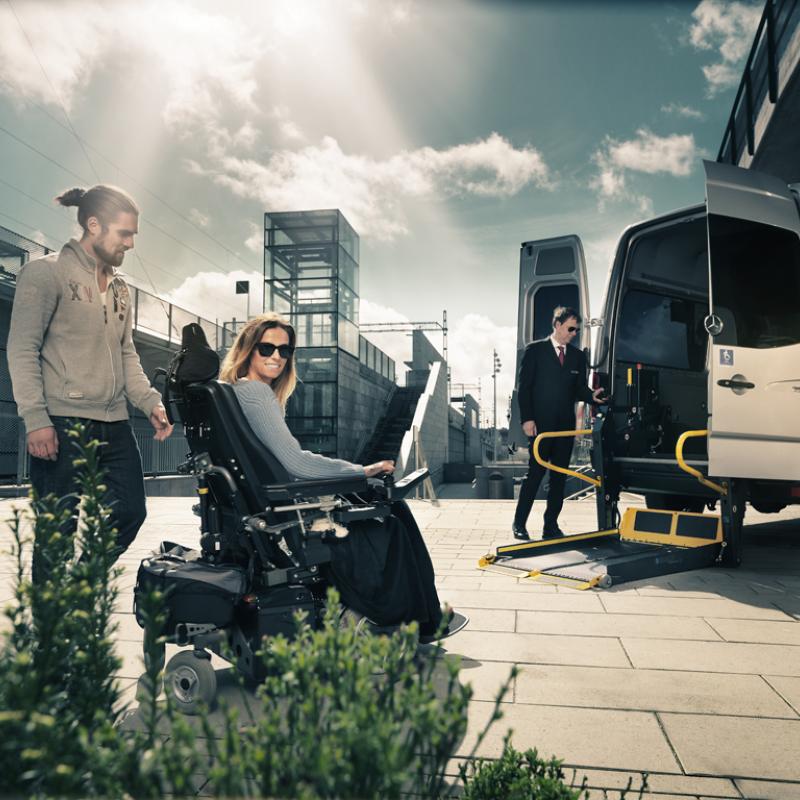 User in powerchair with her carer moving towards a minibus with a deployed wheelchair lift. 